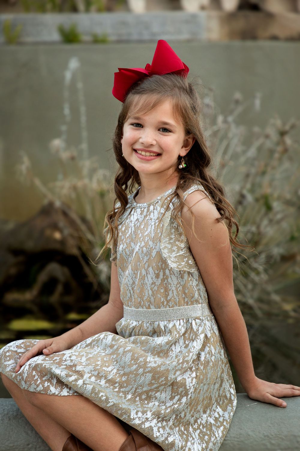 Little girl in golden sequin dress with red bow in her hair sitting on a bench.