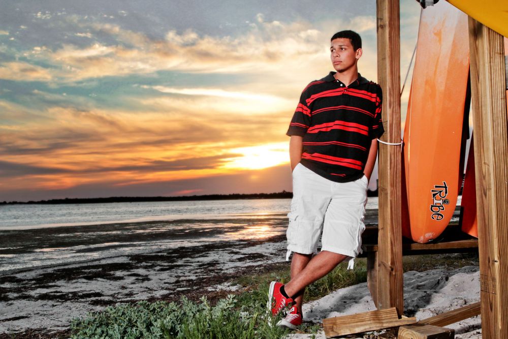 Young man in stripped shirt and tan shorts standing in the sand posing with his hands in his pockets at sunset on the beach.