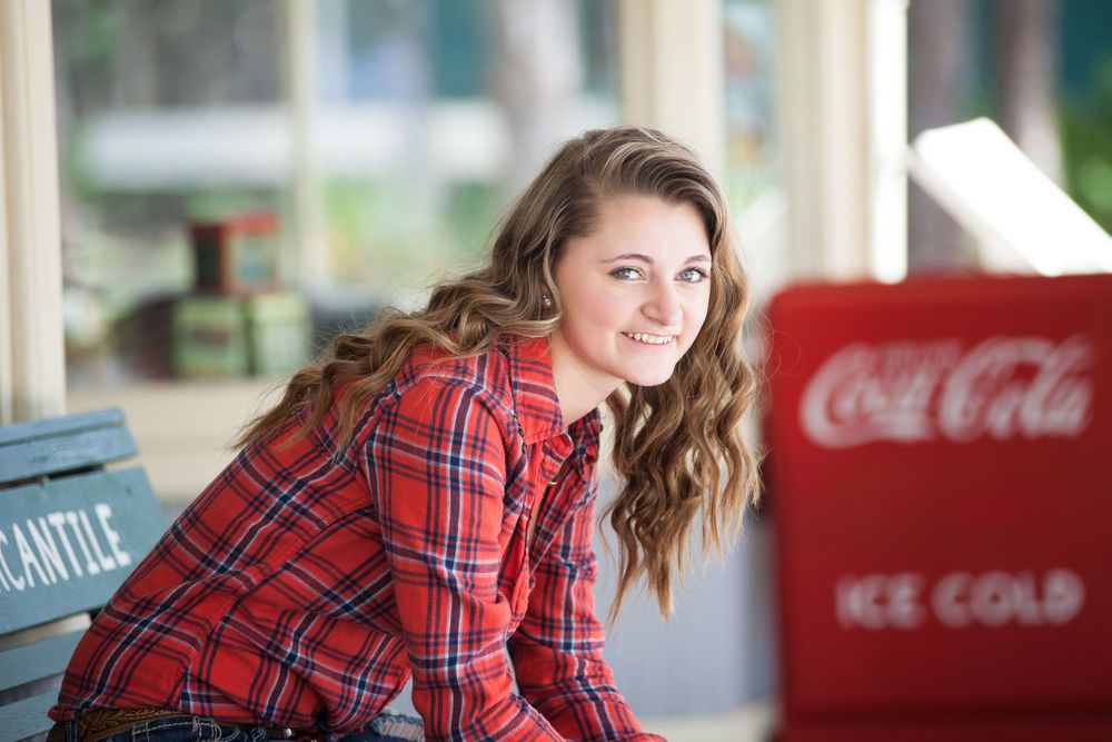 Girl in red plaid shirt sitting on green wooden bench next to a Coca Cola chest.