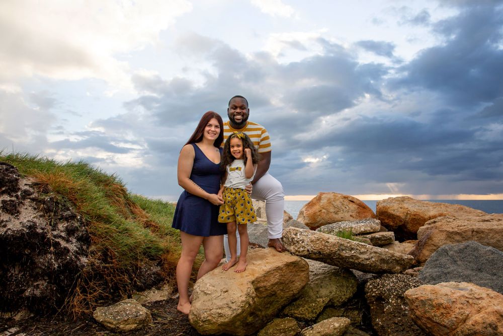 Woman in blue dress with a man in a stripped shirt standing on the rocks with a little girl as the sun is setting.