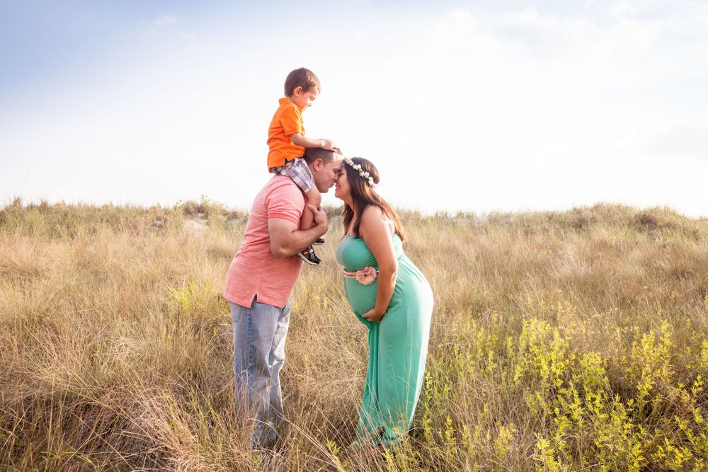 Pregnant woman in a green dress kisses her partner, who carries a young child on his shoulders, in a sunlit grassy field.
