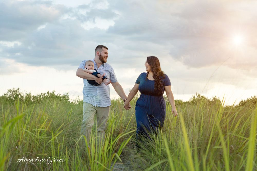 Dad holding a baby while also holding Mom's hand in a field at sunset.