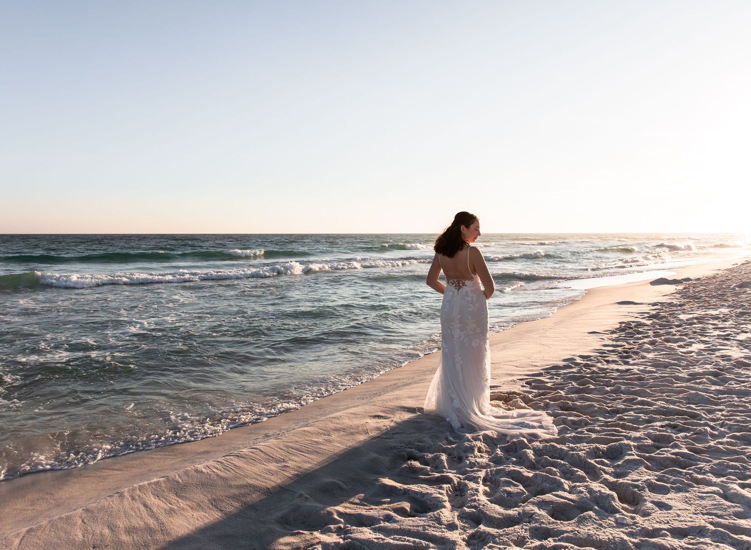 Person in flowing white dress walking along beach shore at sunset.