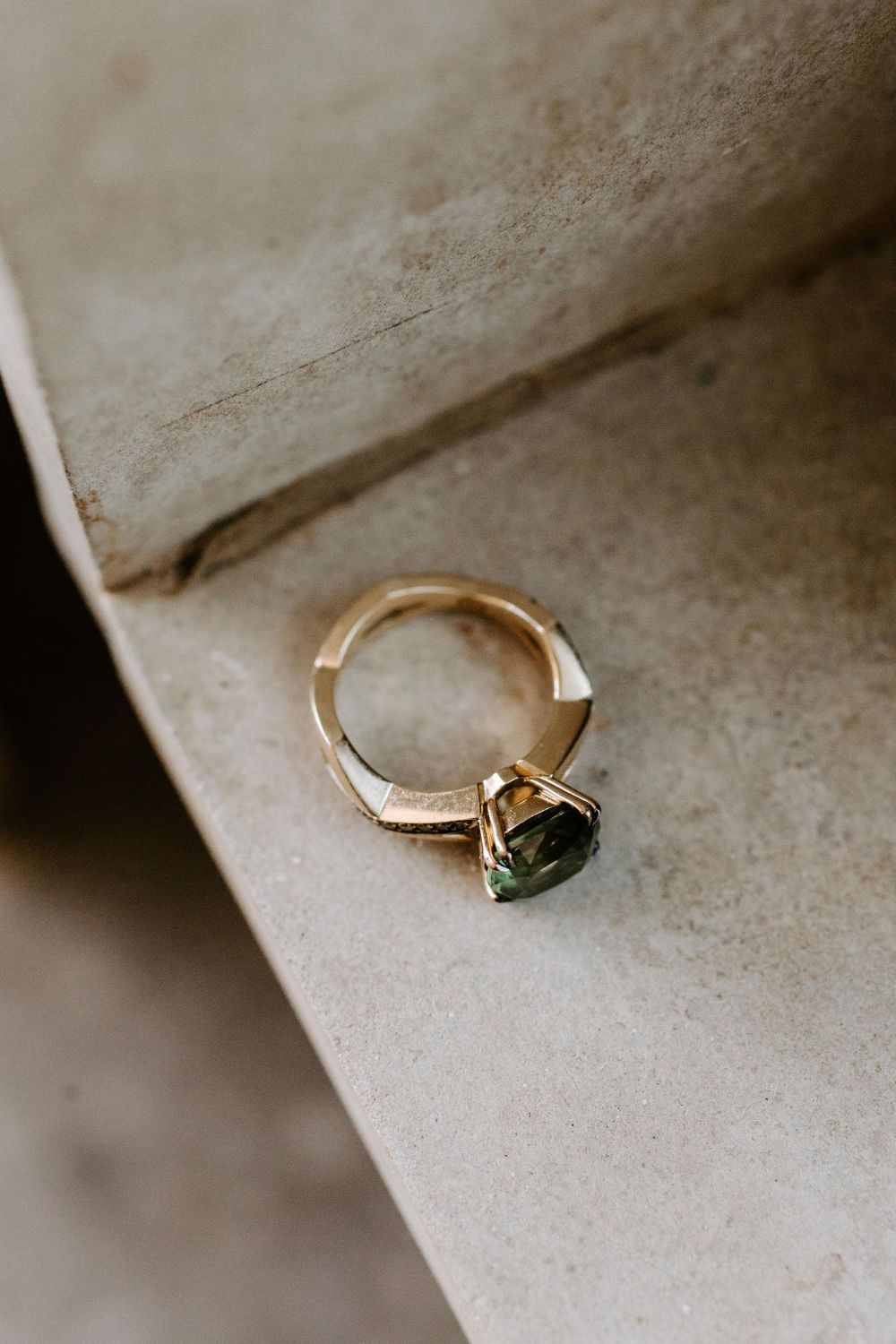 Gold engagement ring with green gemstone sitting on textured surface.
