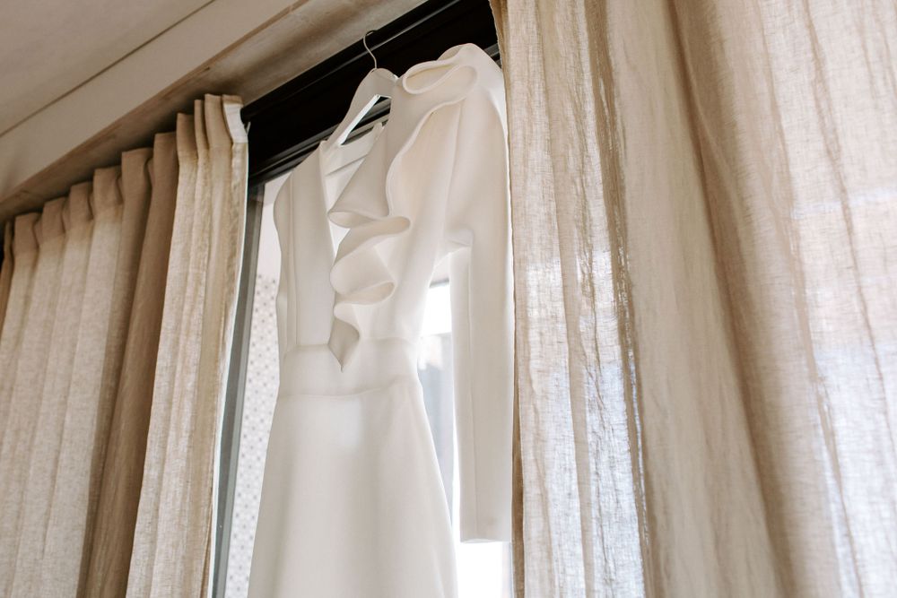 White wedding dress hanging by sheer curtains in natural light.
