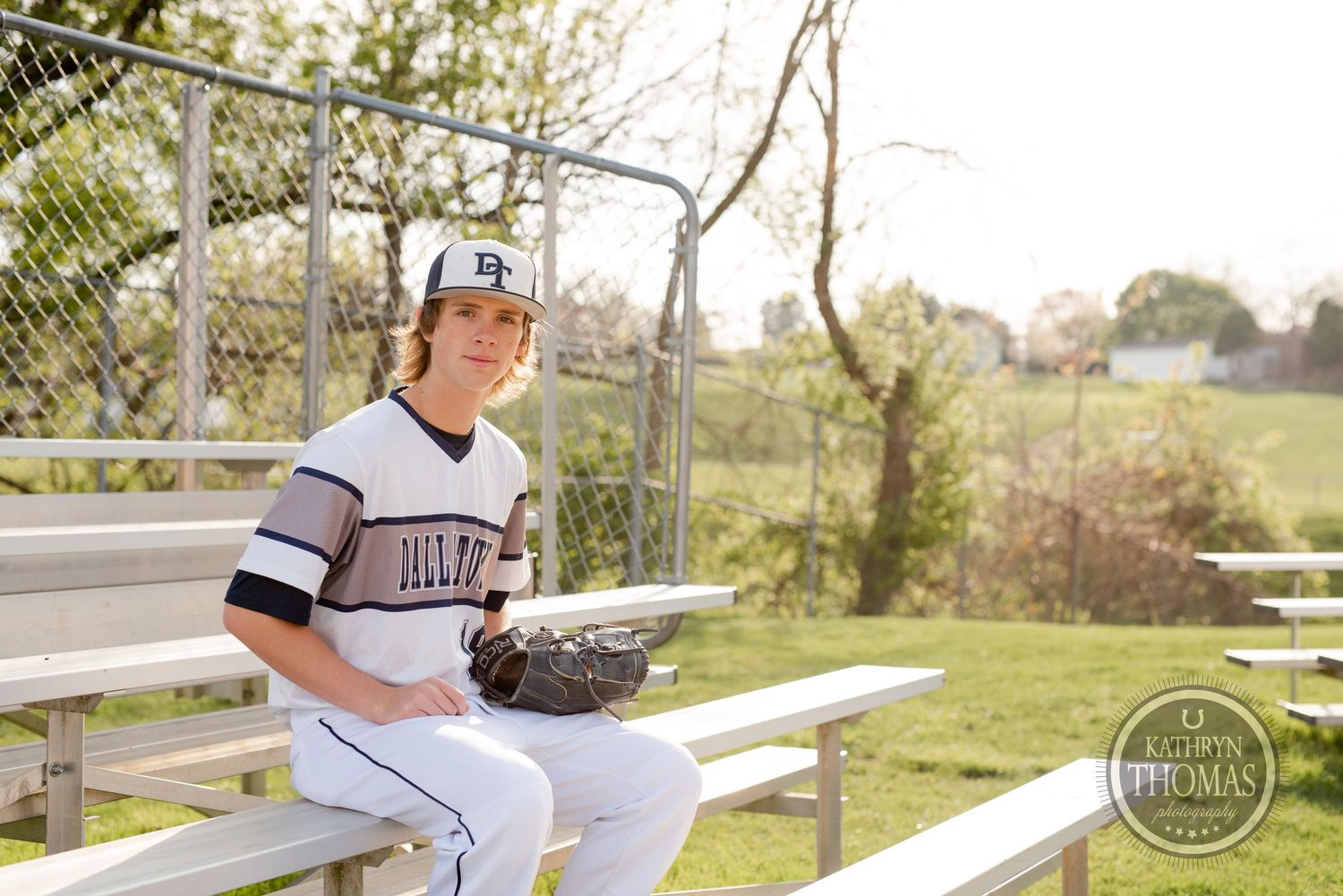 Baseball player in white uniform sits on bleachers near dugout during sunny day.