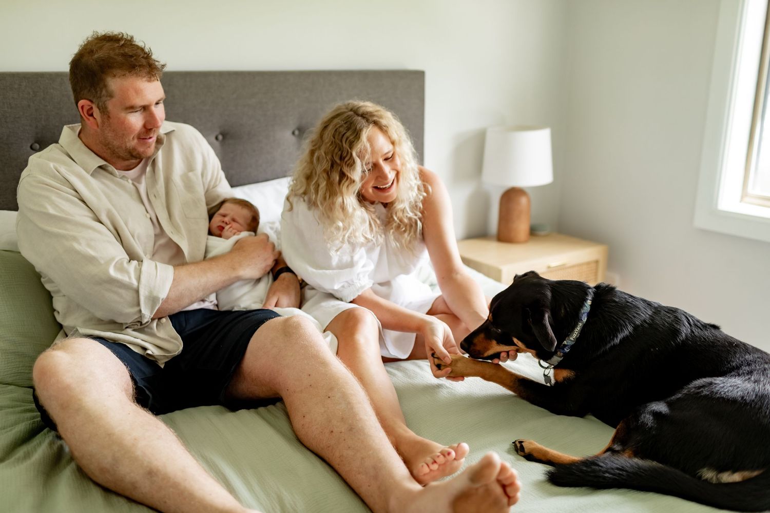 Family relaxing on bed with black dog and newborn baby.