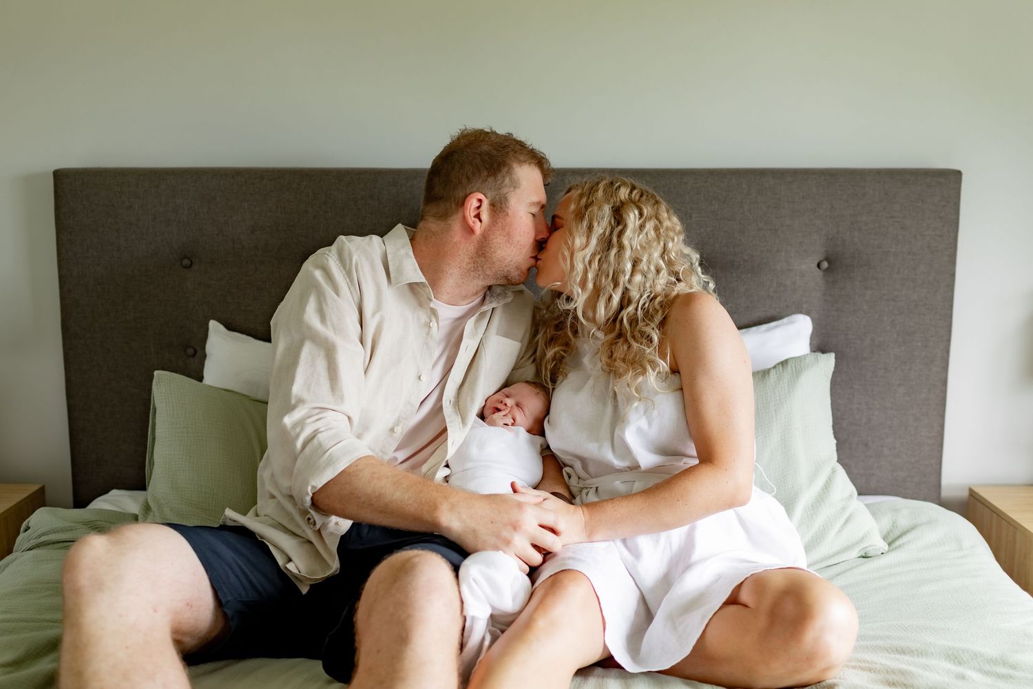 Intimate family moment with newborn baby on bed with gray headboard.