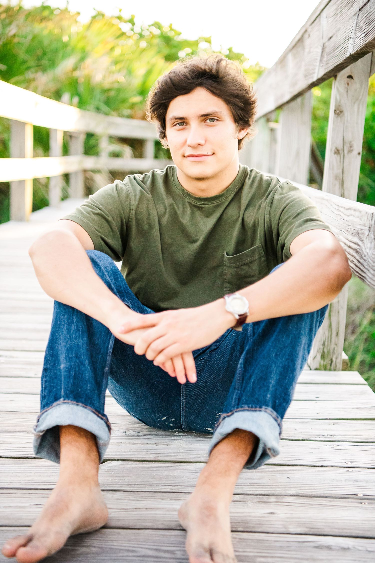 A casual portrait of a person sitting cross-legged on a wooden boardwalk.