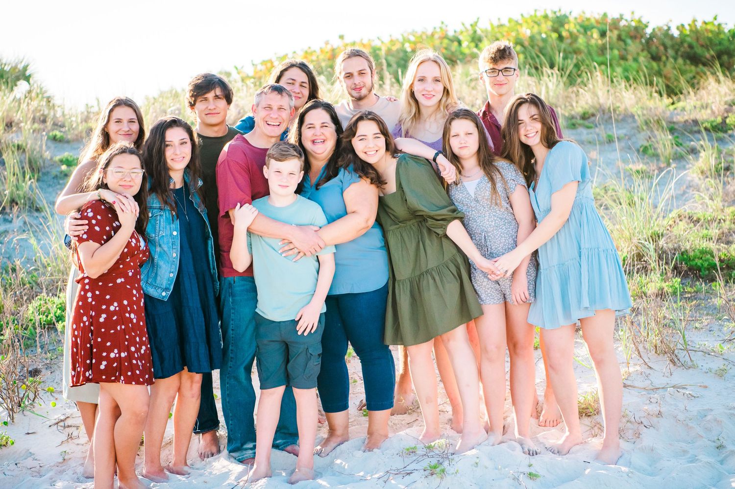 A large group poses together for a casual beach photo in coordinated blue and neutral outfits.