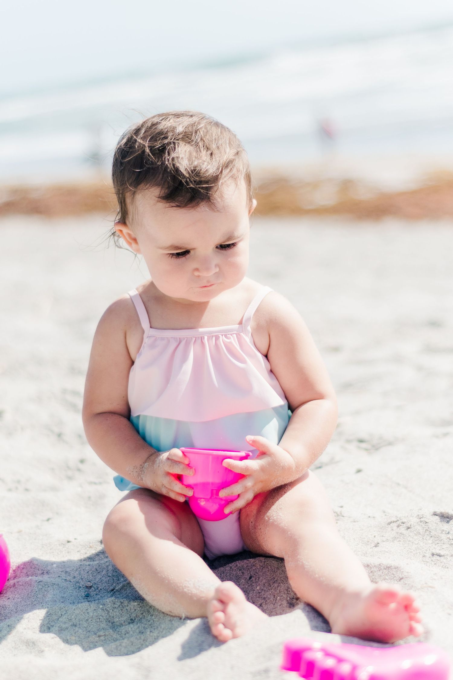 Small child in pink outfit playing with toys while sitting on sandy beach.