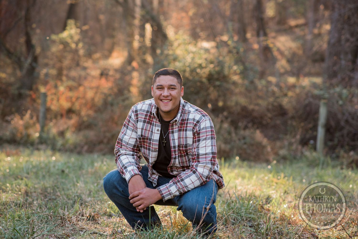 A person in a plaid flannel shirt and jeans sits casually outdoors during golden hour in a wooded setting.