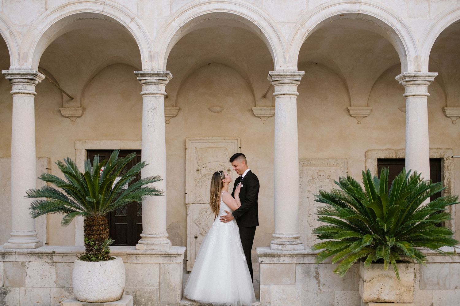 A wedding couple stands between elegant stone arches and potted palm plants at a Mediterranean-style venue.