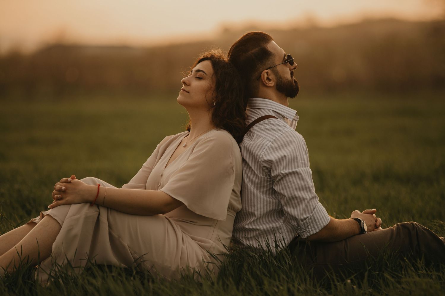 A couple sits back-to-back in a grassy meadow during golden hour.