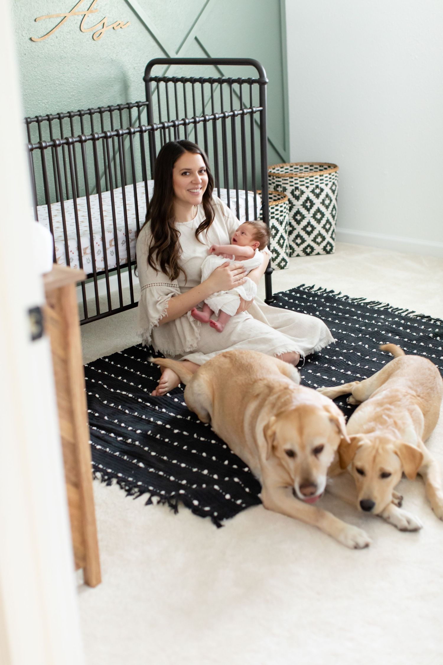 Nursery scene with two golden retrievers lying beside woman and baby on patterned rug.
