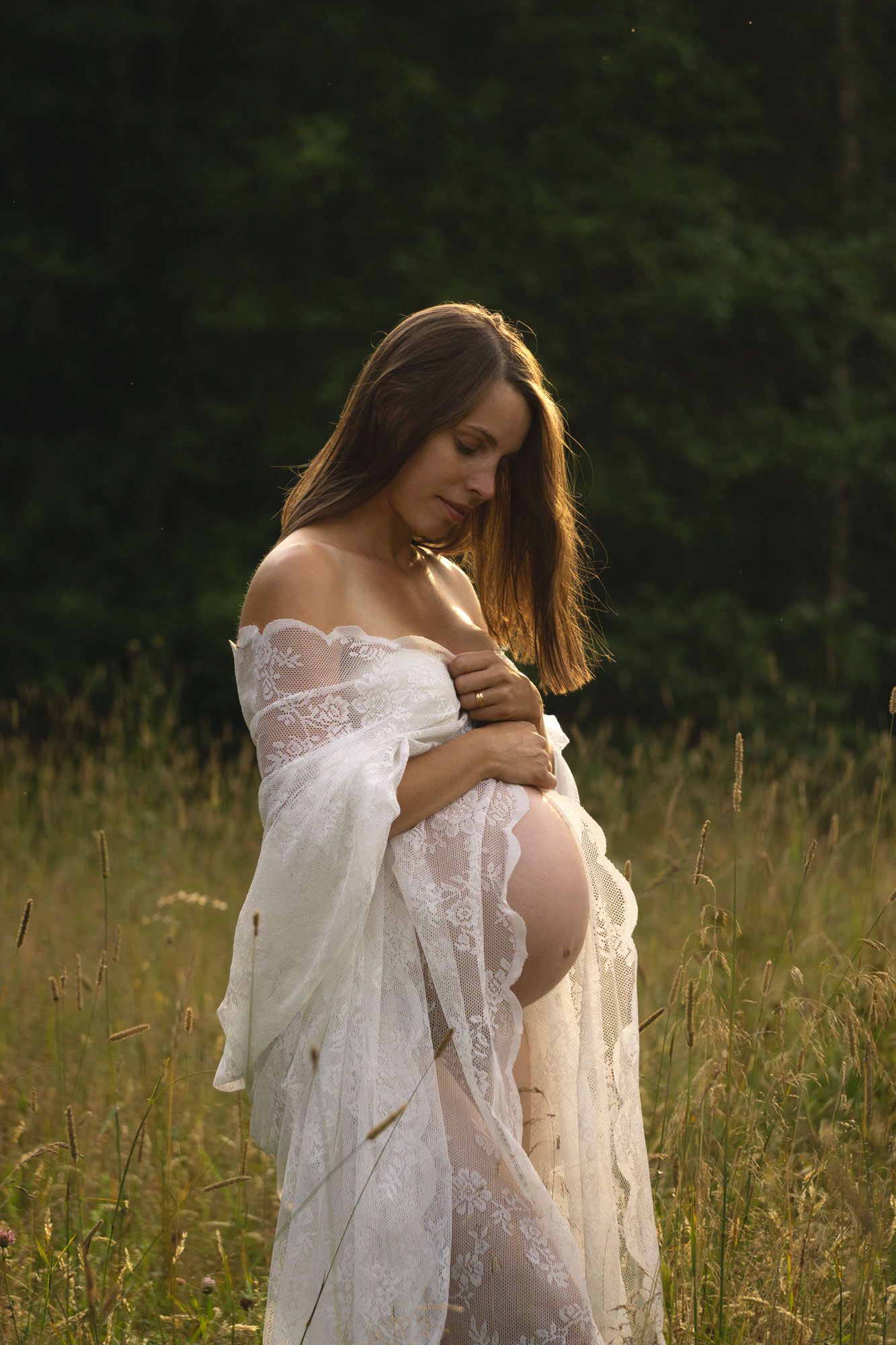 En gravid kvinna med virkad gardin om sig håller händerna ovanför gravidmagen. Hon står i högt ängsgräs. Det är en solig kväll.