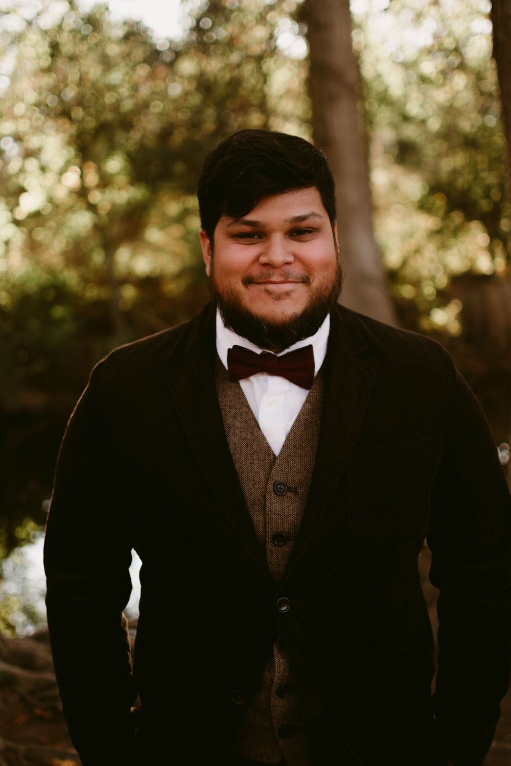 Formal portrait in classic black tuxedo and bowtie with brown vest in forest setting.