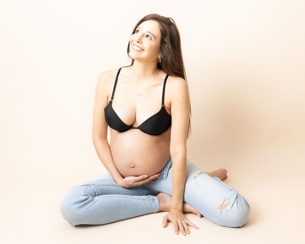 An expectant mother in blue jeans and black bra sits in relaxed pose while gazing upward against beige backdrop.