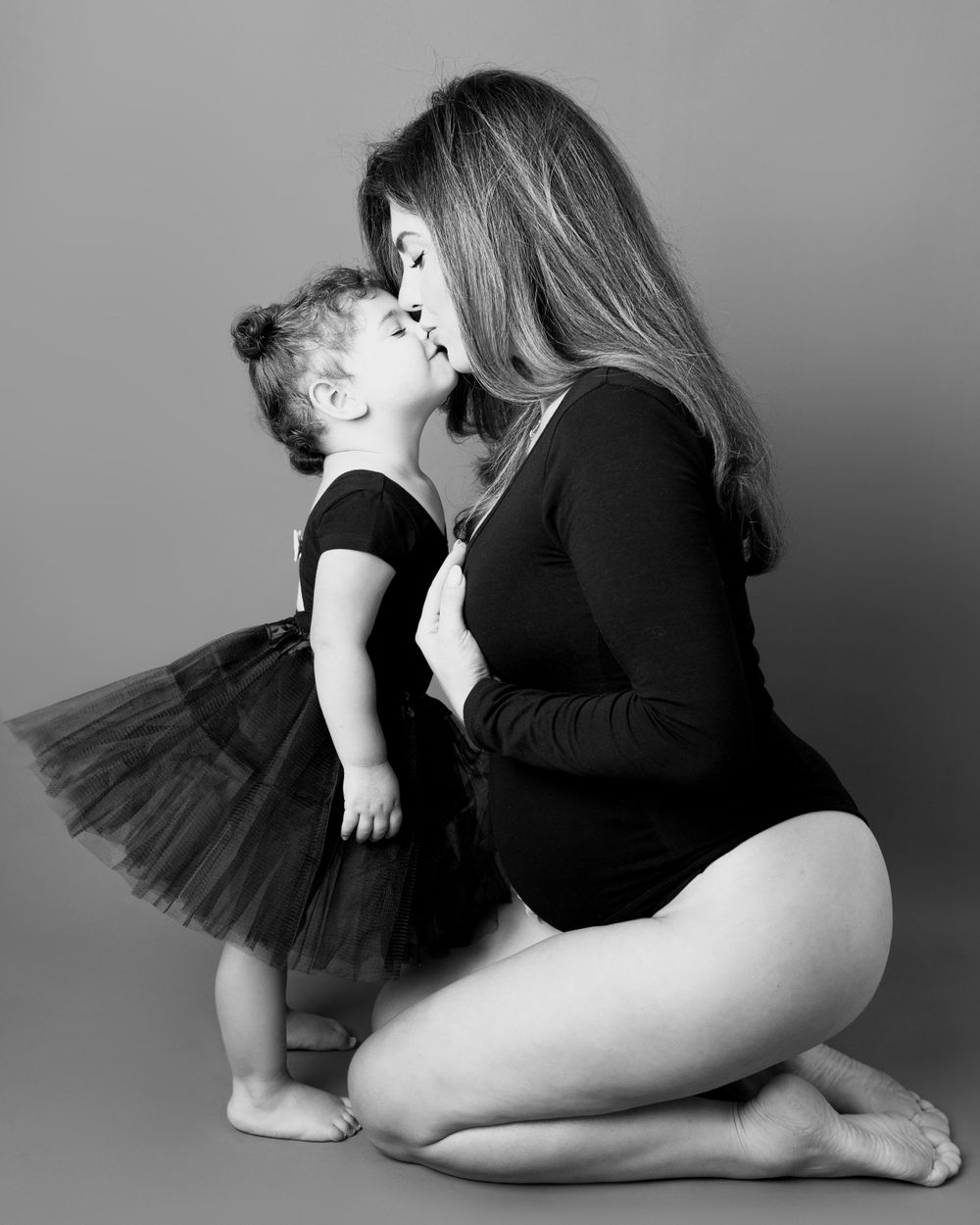 Black and white portrait of tender embrace between adult wearing black bodysuit and small child in black tutu.
