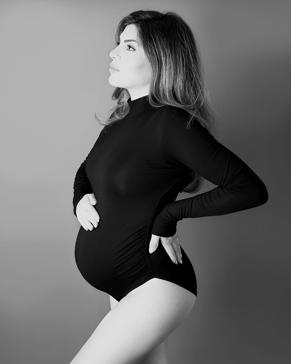 A black and white artistic maternity photo showing an expectant mother in a black bodysuit posing in profile view.