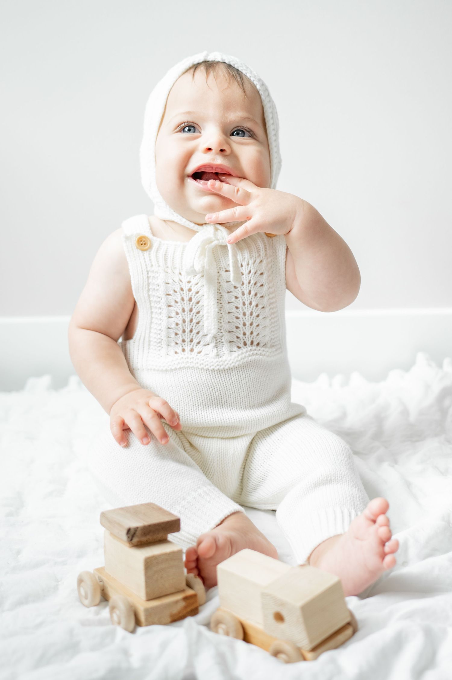 little boy in white knit outfit smiles up at camera with hand in his mouth and wooden toys around him