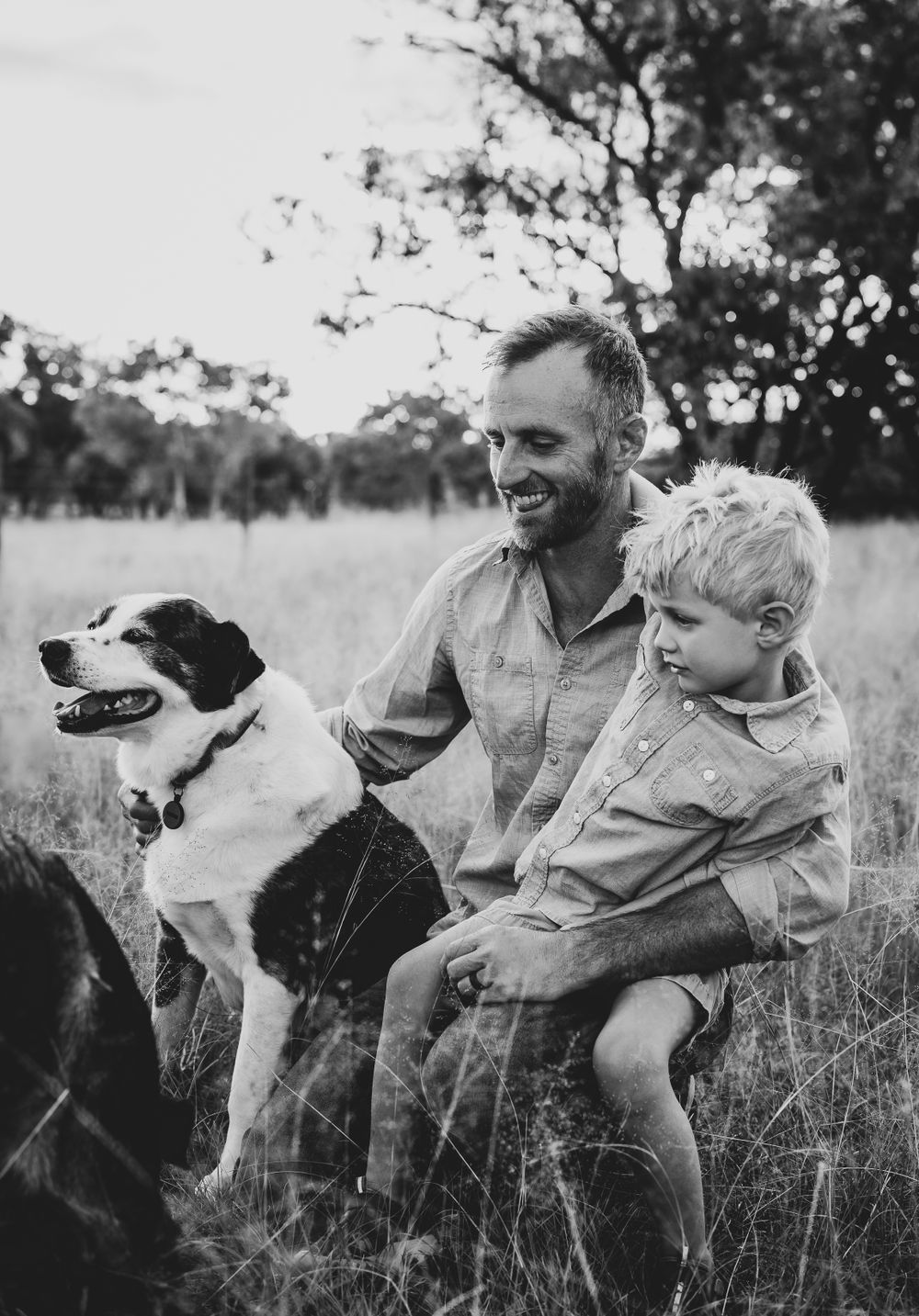 Black and white image of parent and child sitting with their dog in rustic field setting.