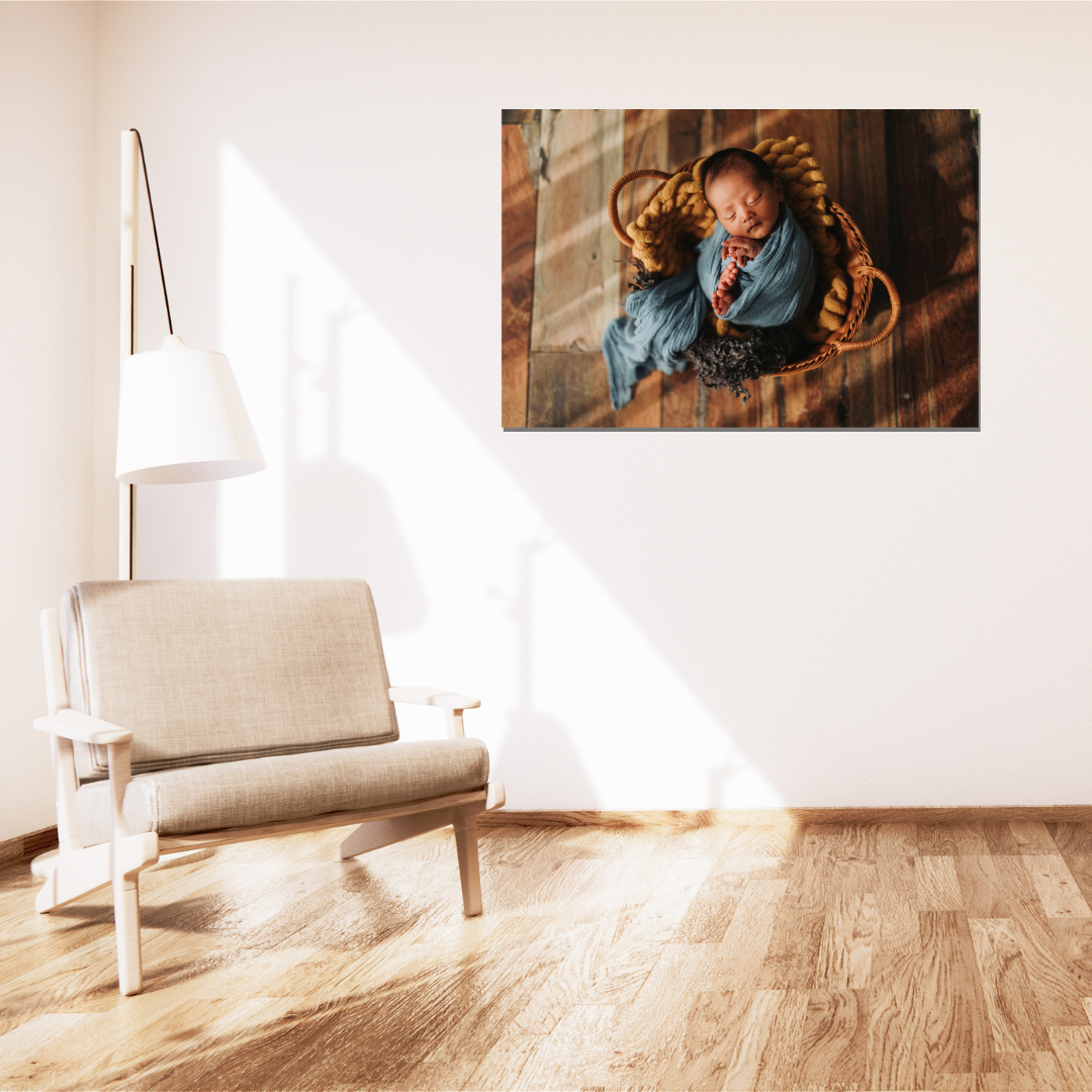 Canvas wall art of a swaddled newborn in blue, sleeping in a wicker basket, displayed above a modern chair.