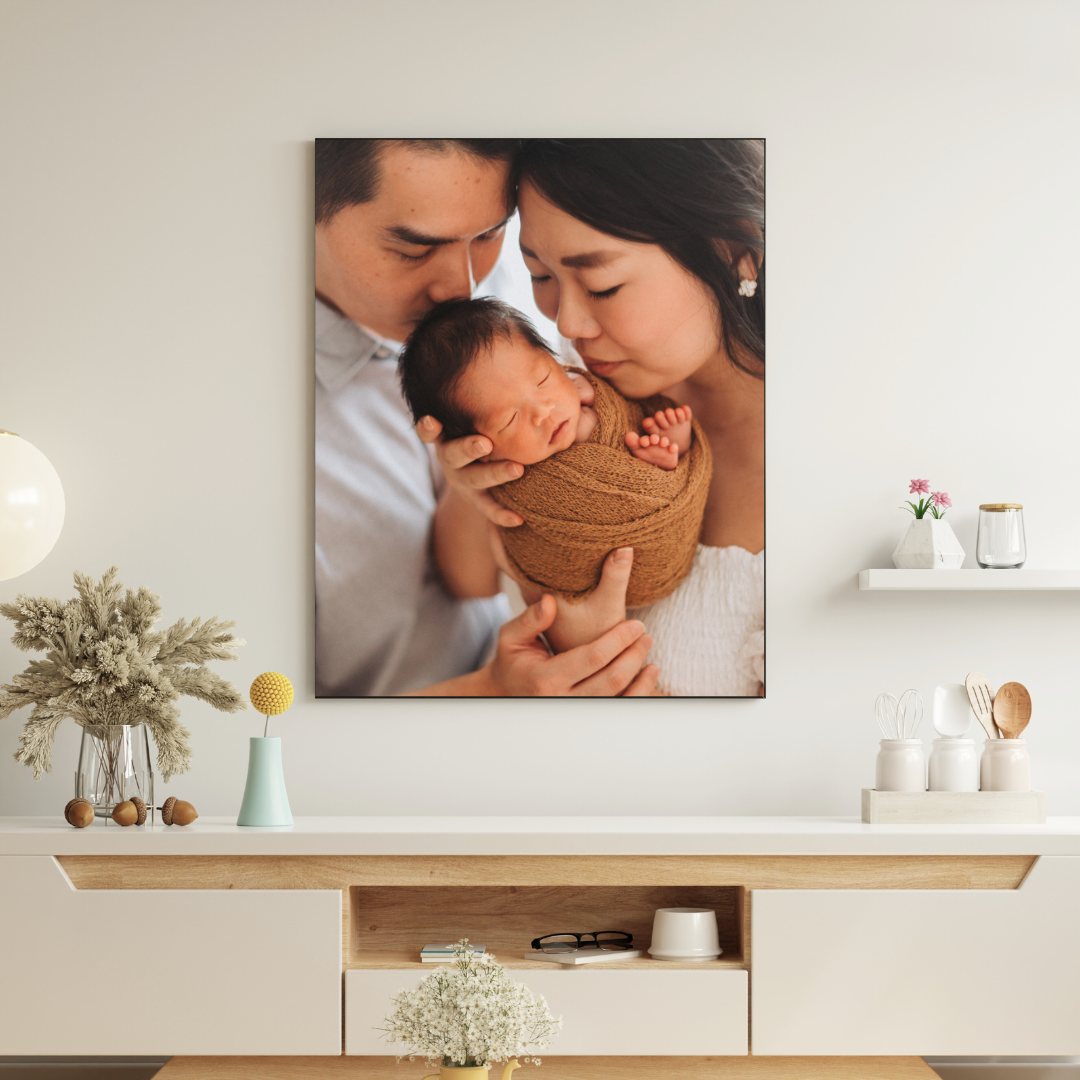Canvas print of parents tenderly kissing their swaddled newborn baby, displayed above a modern white sideboard.