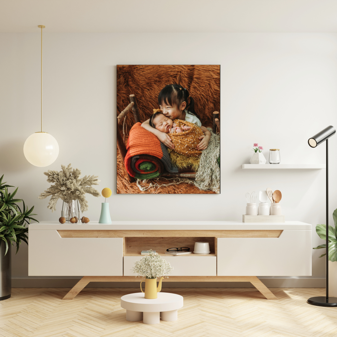 Canvas wall art of a young child lovingly embracing a sleeping newborn in a rustic setting, hung in a bright room.