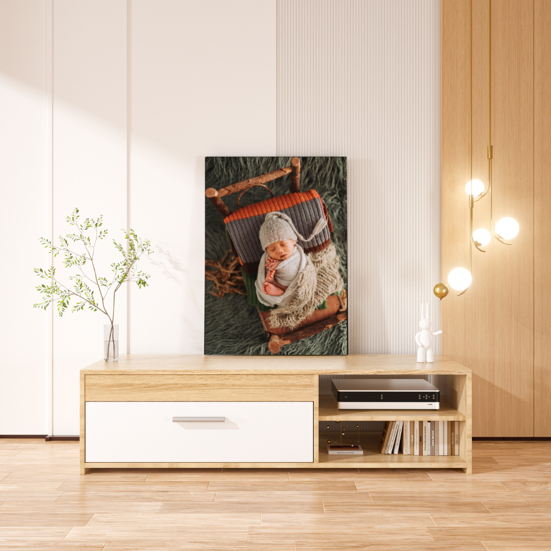 Canvas wall art of a sleeping newborn in a rustic wooden sled, leaning against a TV stand in a bright living room.