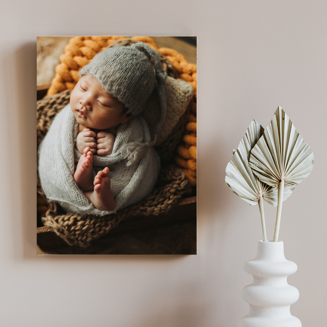 Canvas wall art of a sleeping newborn in a gray knit wrap and hat, displayed on a wall beside a decorative vase.
