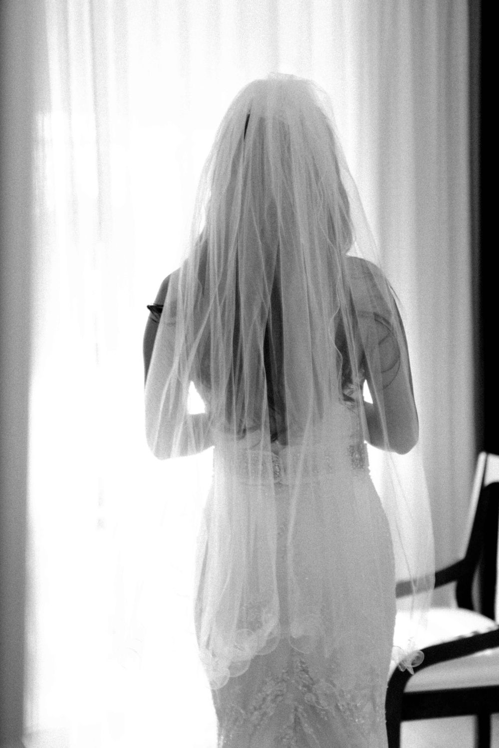 Black and white silhouette of a bride in a white wedding gown and veil standing near a window.