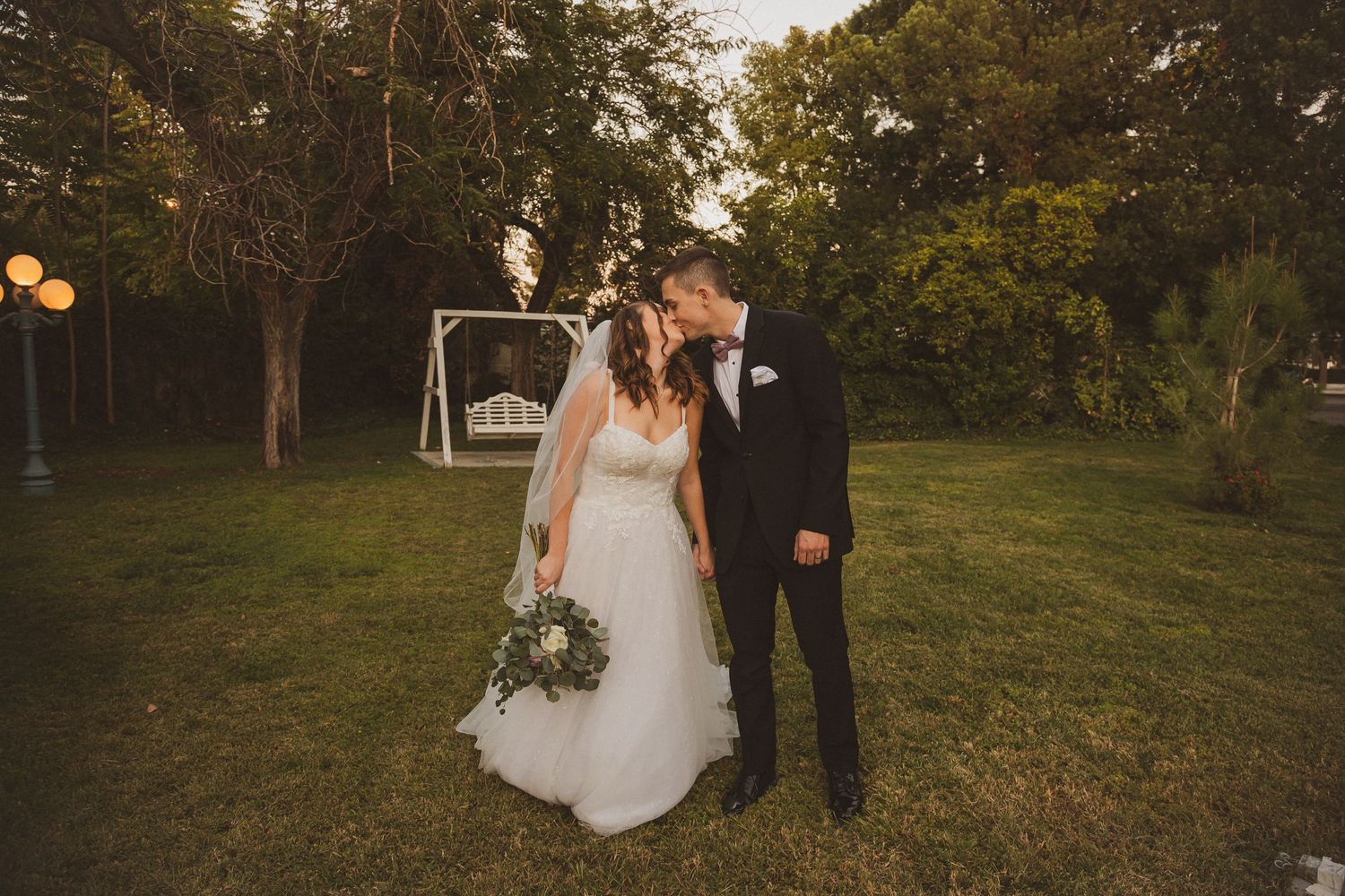 A couple poses together in a garden setting during golden hour with string lights in the background.