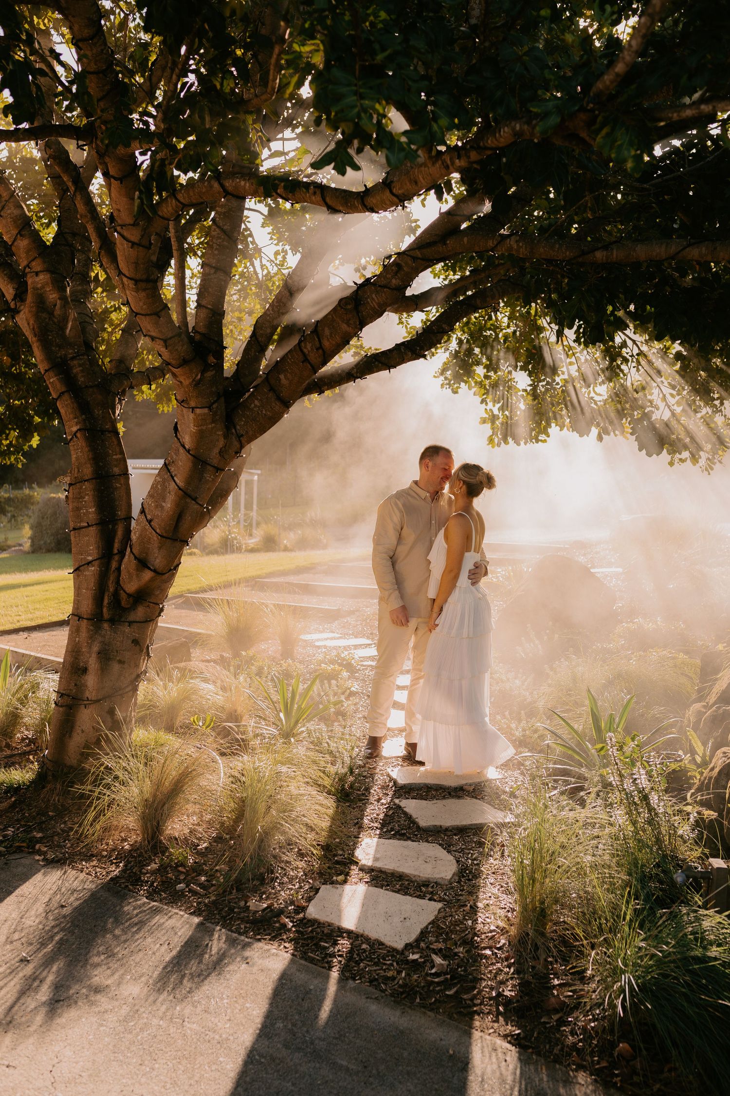 Romantic couple embracing on stone steps under dreamy sunlit tree canopy.