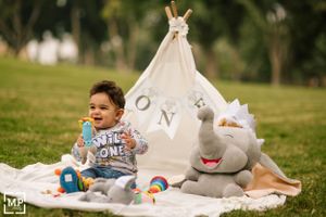 Baby sits on blanket with toys near white teepee tent and plush animals.