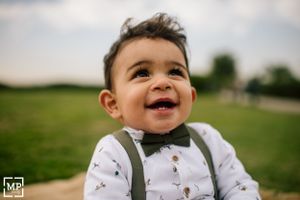 A toddler in suspenders and bow tie smiles brightly outdoors.