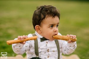 A young child in suspenders playfully bites a breadstick outdoors.