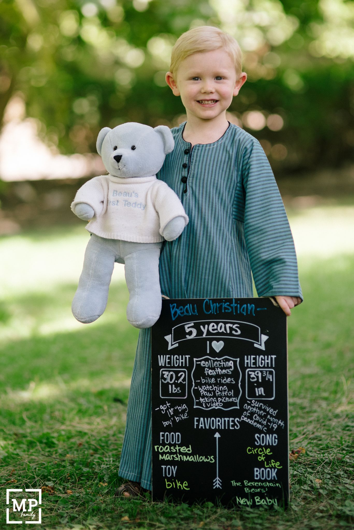 Child holding teddy bear poses with milestone chalkboard showing 5 year achievements.