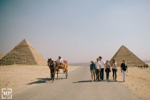 Tourists walking near horse-drawn carriage with two pyramids visible in desert background.