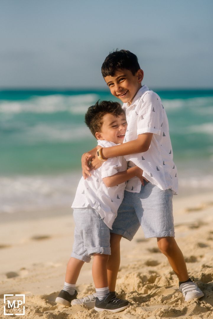 Two boys in matching white outfits share a joyful embrace on a sunny beach with turquoise waters in the background.