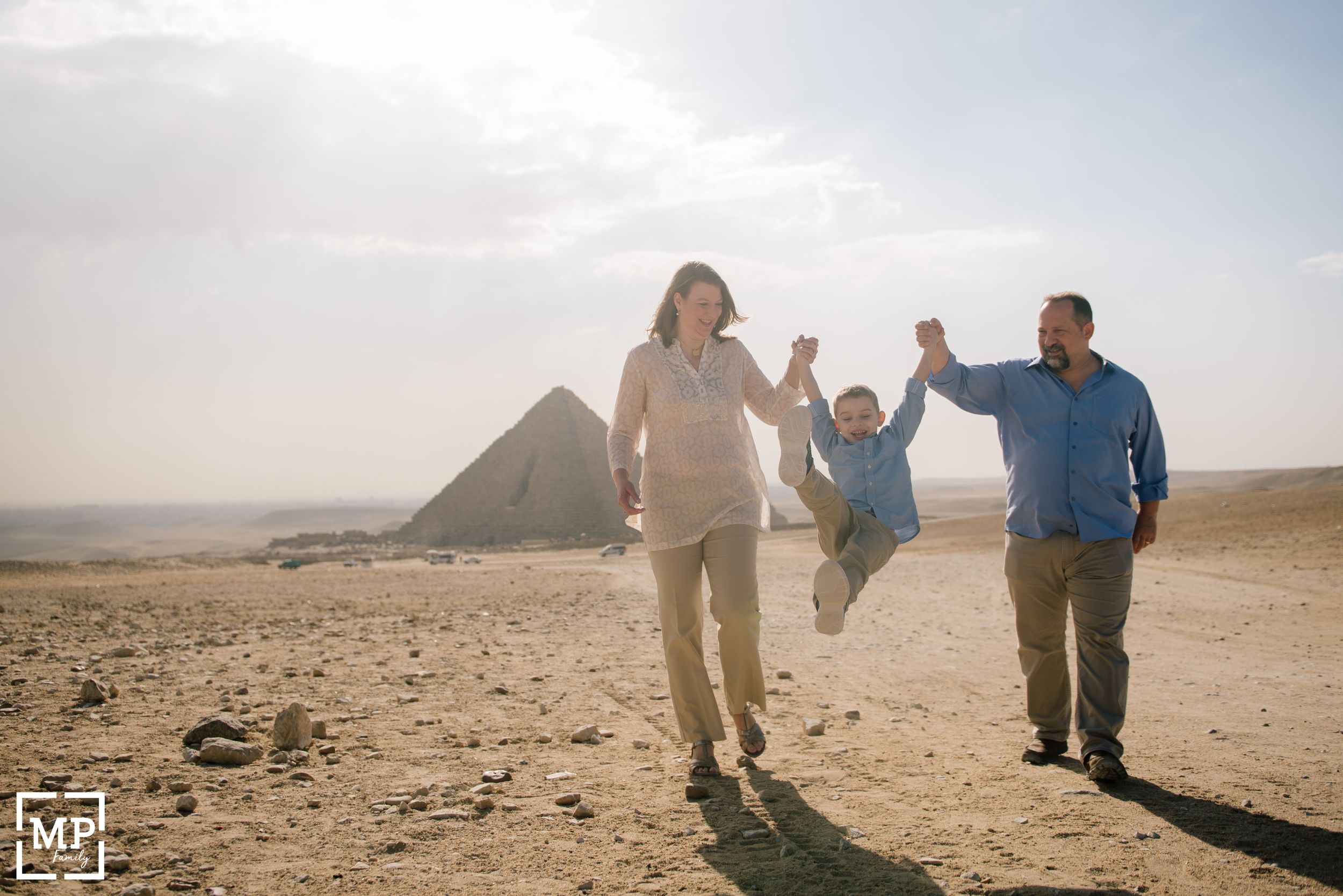 Family swings child while walking near the ancient pyramids of Egypt in the desert landscape.