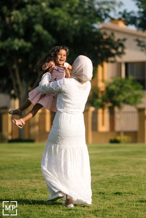 Parent lifting and playing with child in white dress on sunny green lawn.