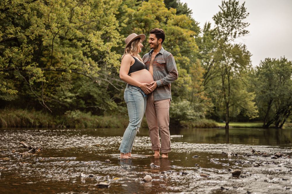 A couple stands in shallow flowing water near a tree-lined riverbank during a photo session.
