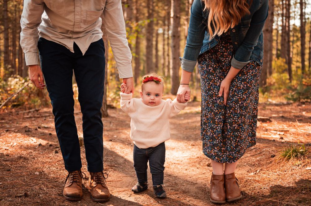 A small child takes first steps on a forest trail while being supported by adults on both sides.