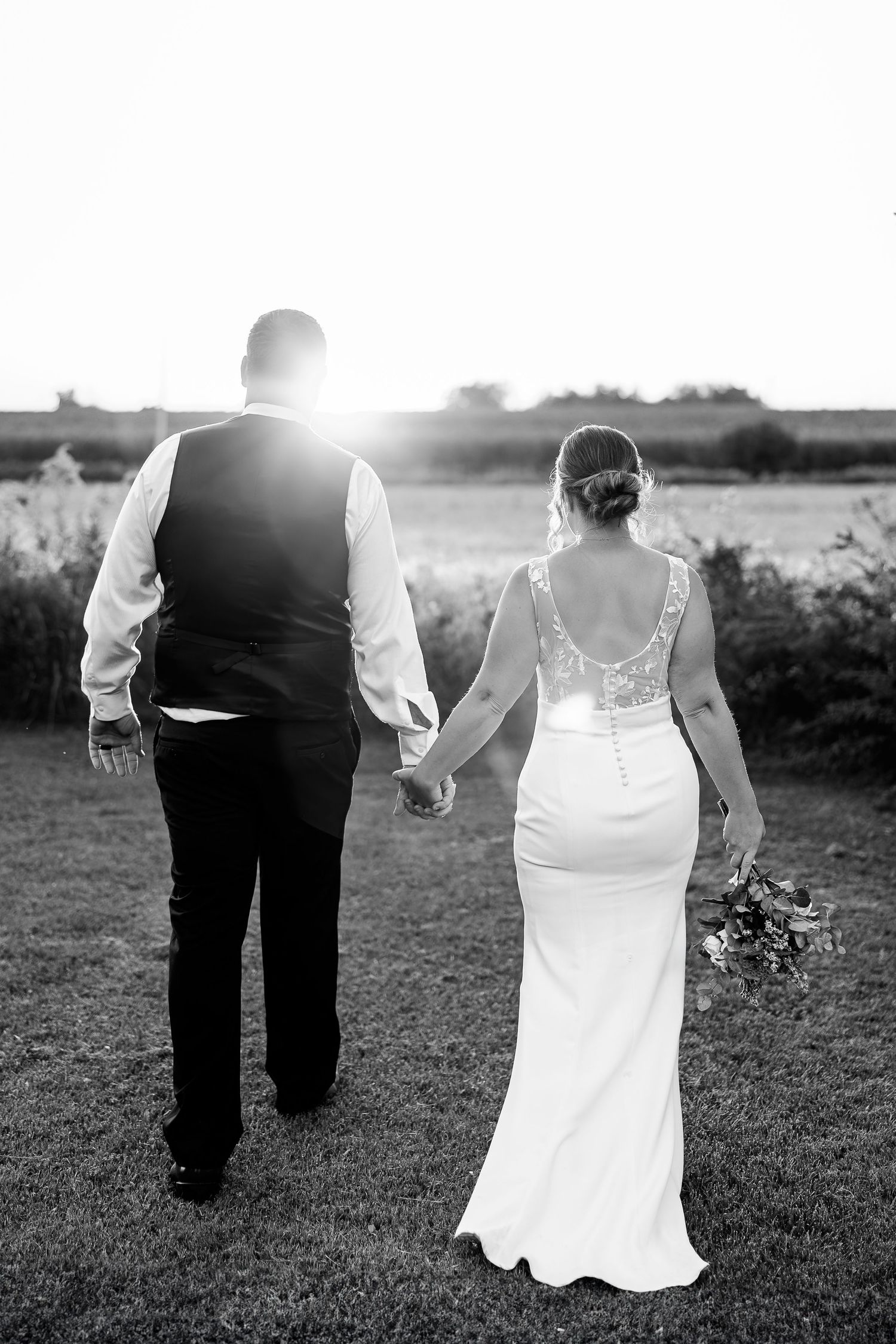 Silhouetted couple walking hand in hand at sunset, with bride in elegant lace-back dress.