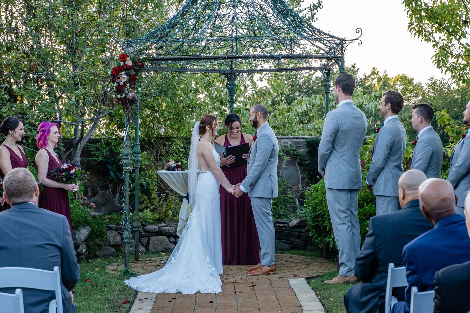Outdoor garden wedding ceremony under a green gazebo with groomsmen and guests in attendance.