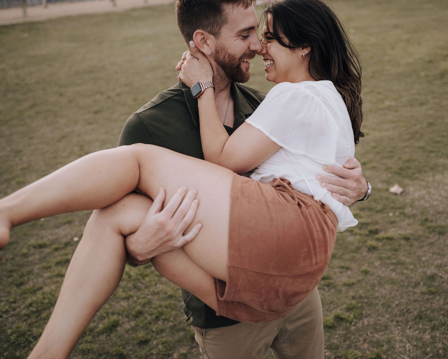 A romantic couple shares an intimate moment as he carries her while standing in a grassy field.