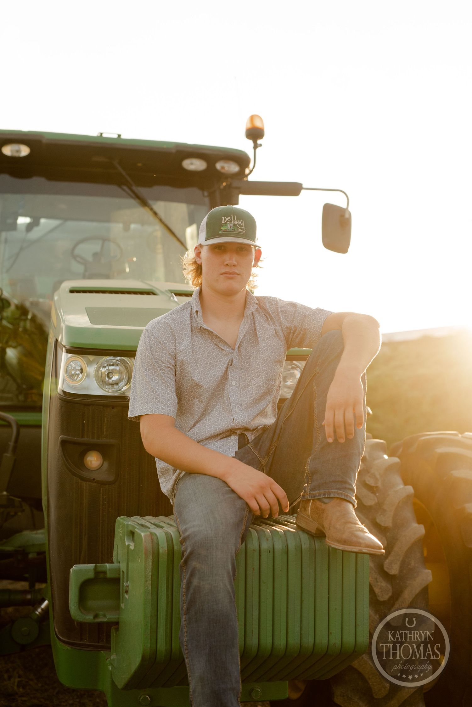 Golden hour portrait of a young farmer sitting on a green John Deere tractor in casual work attire.