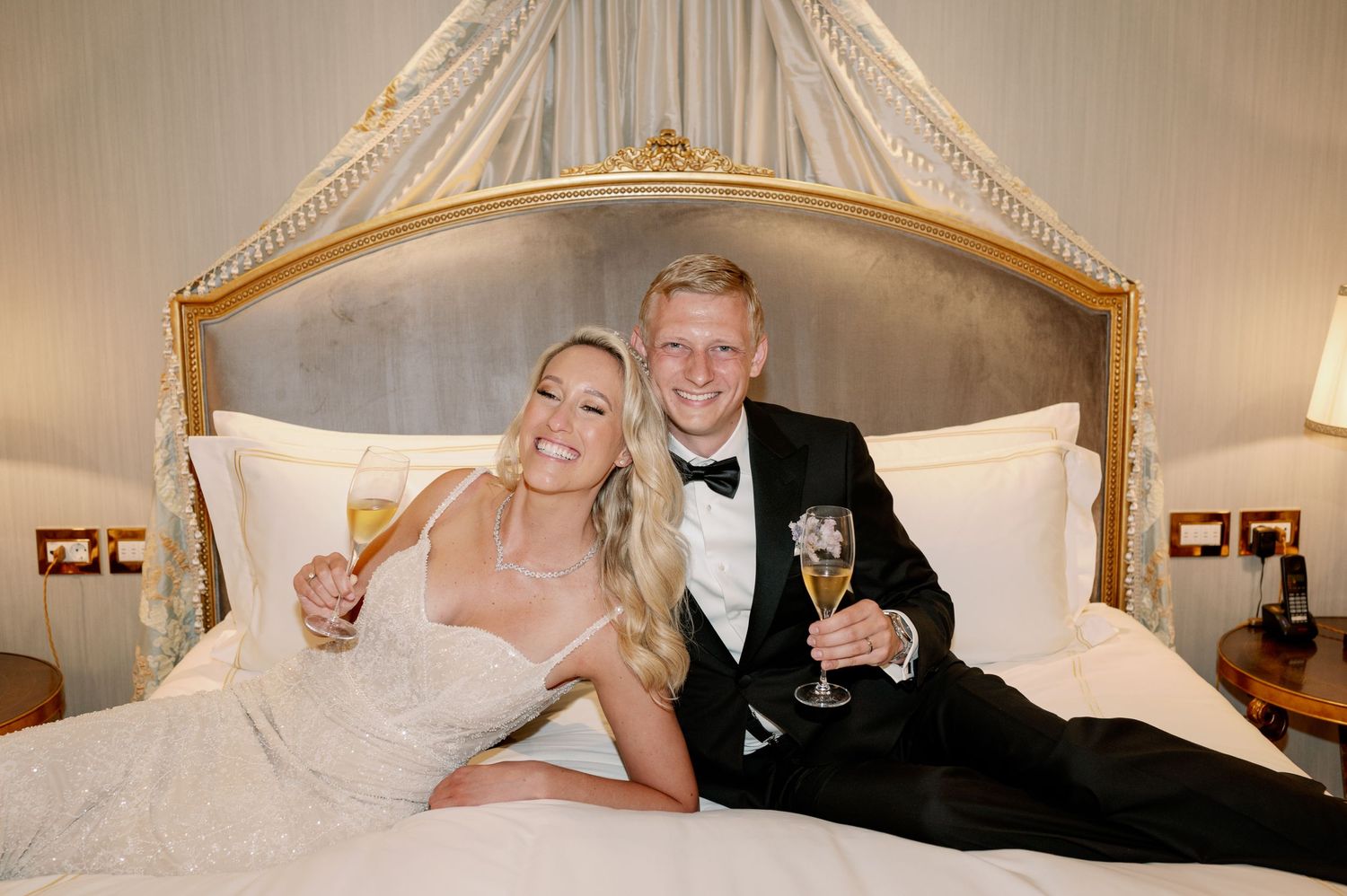 Wedding couple poses with champagne glasses on elegant canopy bed.