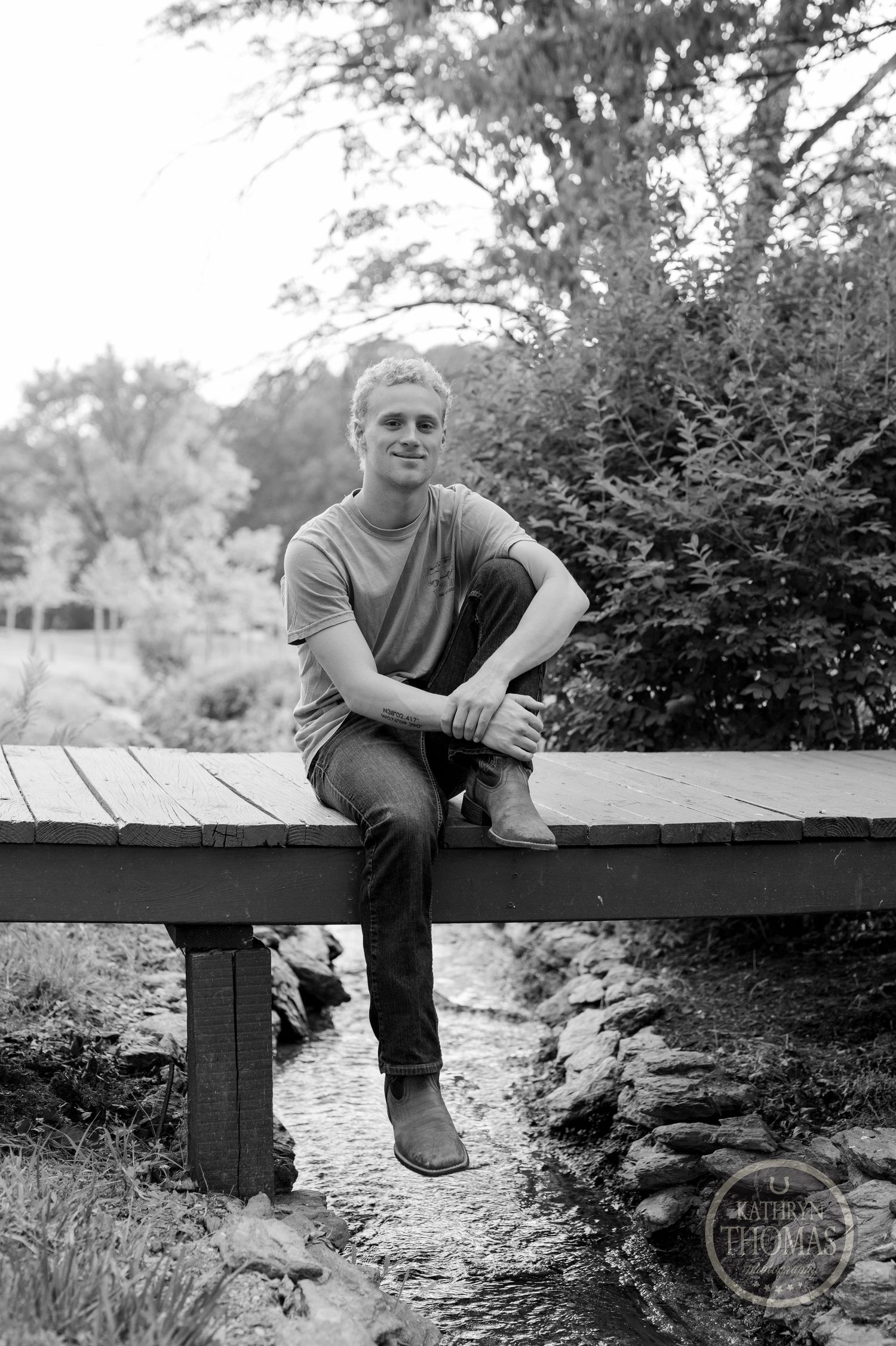 Black and white portrait sitting on wooden bridge over small stream with lush foliage.